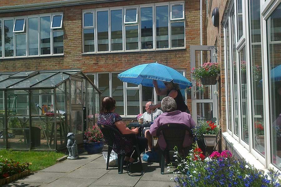 Four people at a garden table next to a greenhouse, one person is putting up a blue parasol