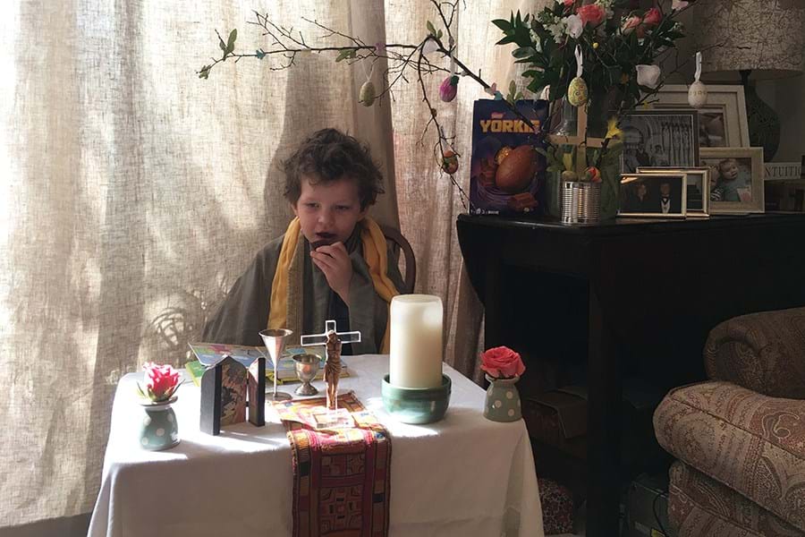 Child dressed as priest standing behind a table with various religious items 