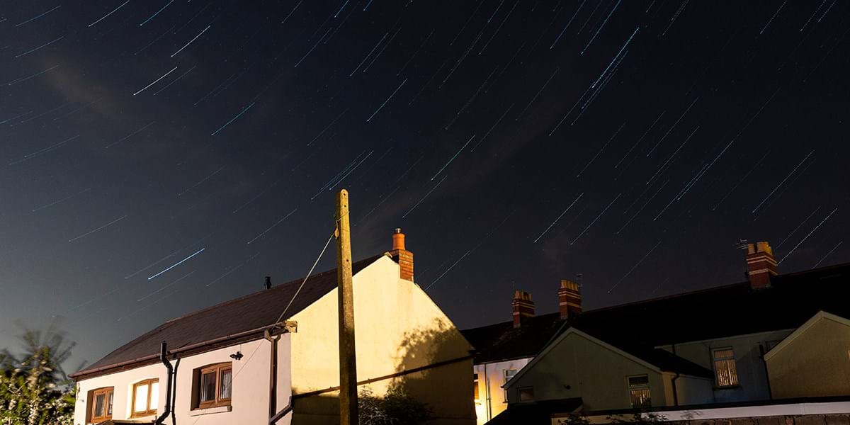 A trail of stars in a dark sky, over the roof of a house