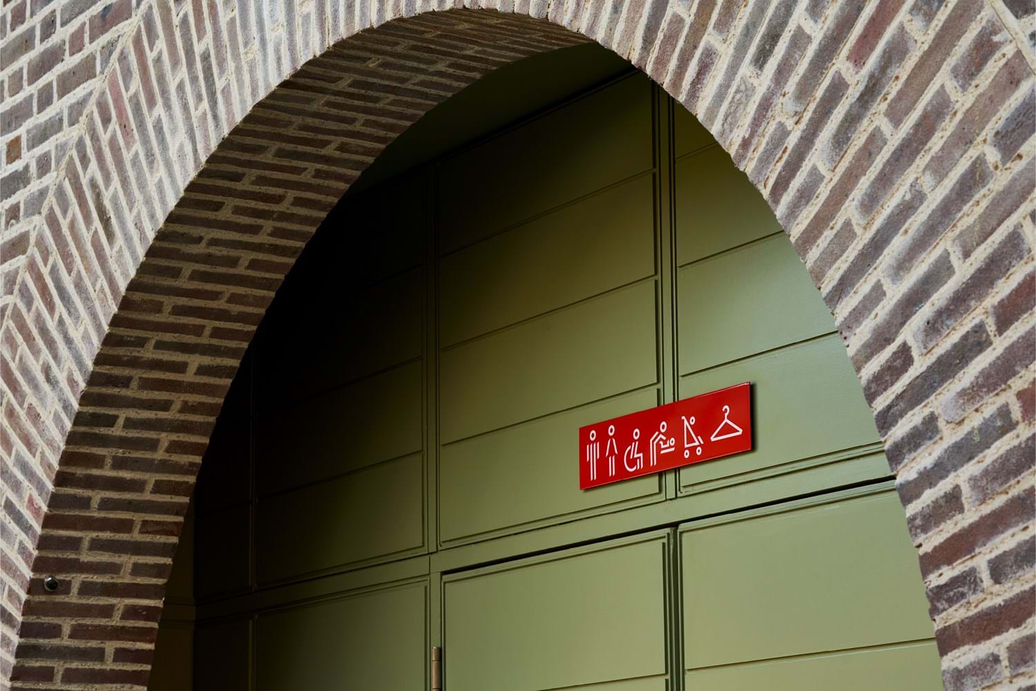 A brick arch framing green doors with red signage, displaying symbols for bathrooms, wheelchair users, changing facilities and buggies.