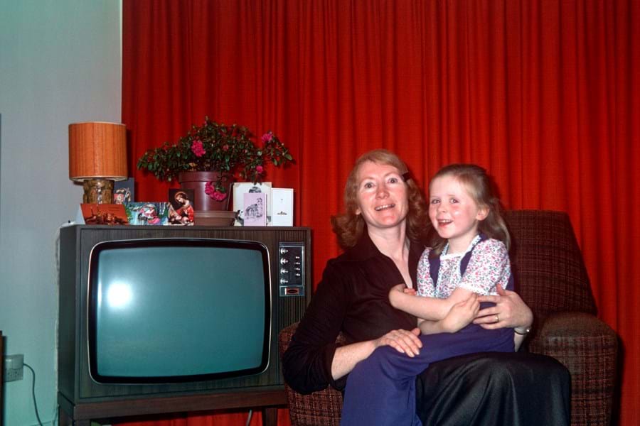 A child sat on a person's lap, next to a television set with plants and a lamp on top