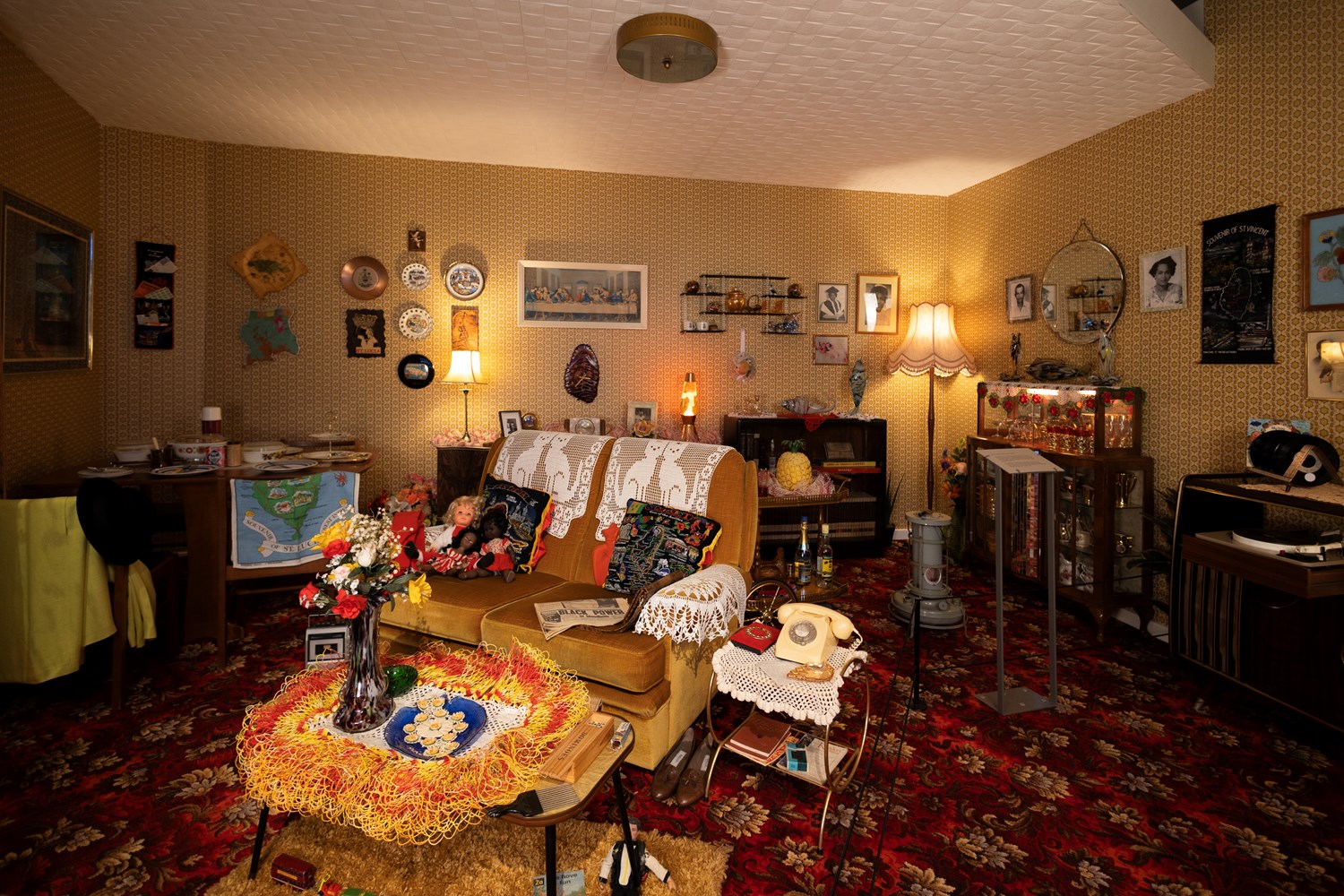 A seventies living room with a mustard yellow sofa in the centre, red patterned carpet and clashing patterns on the walls.