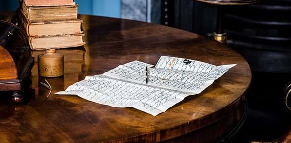 A wooden table with a pile of books in the background and in the foreground a half-written letter and an ink pot