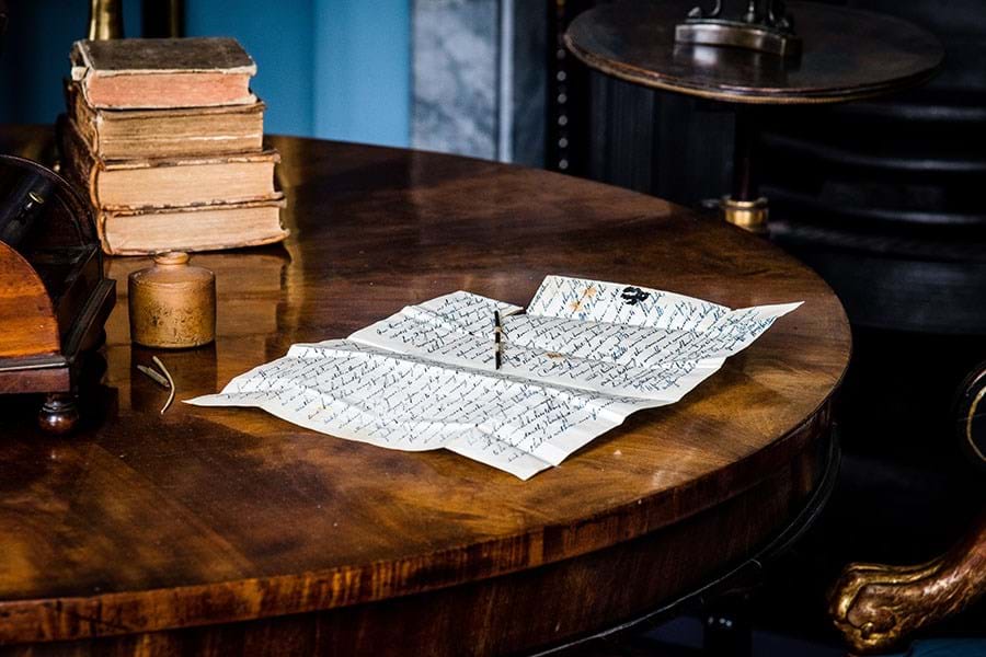 A wooden table with a pile of books in the background and in the foreground a half-written letter and an ink pot