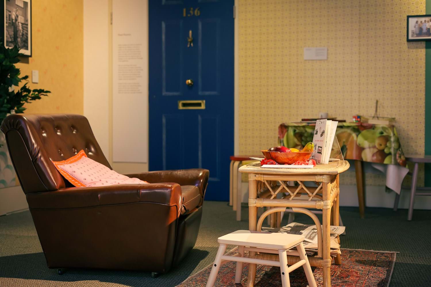 A room set with a dark brown leather lounge seat and a rattan table with a bowl of fruits atop. Behind, yellow wallpaper and a dark blue door with a letter slot in the background.