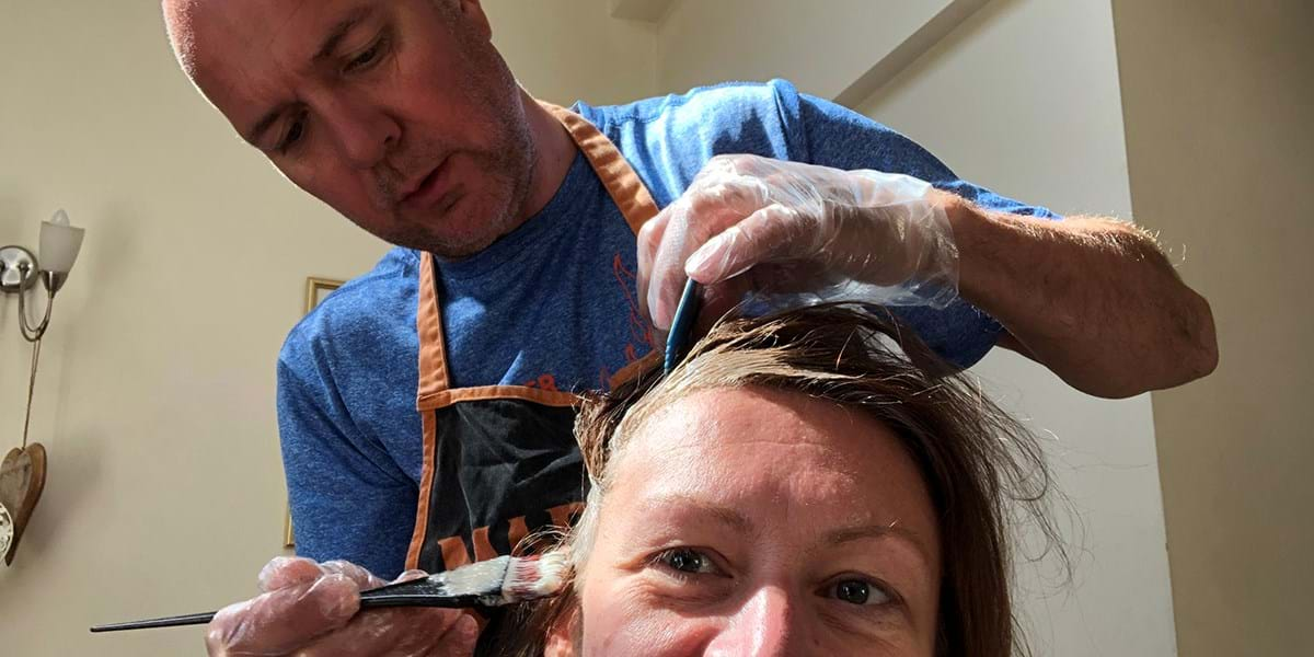 A standing person applying hair dye to a seated person, using a brush 