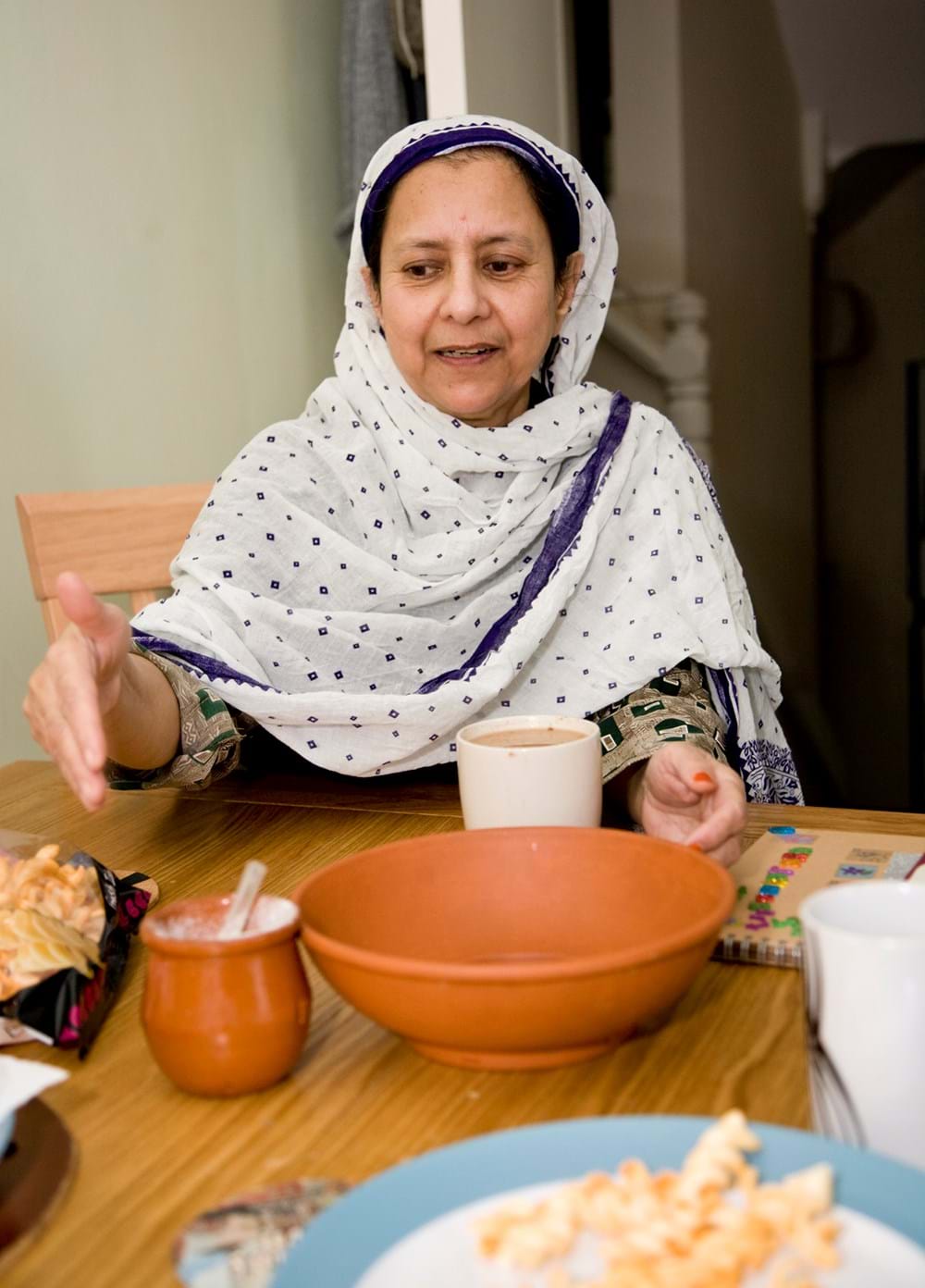 A person wearing a headscarf sitting at a table holding a cup of tea and gesturing with one arm