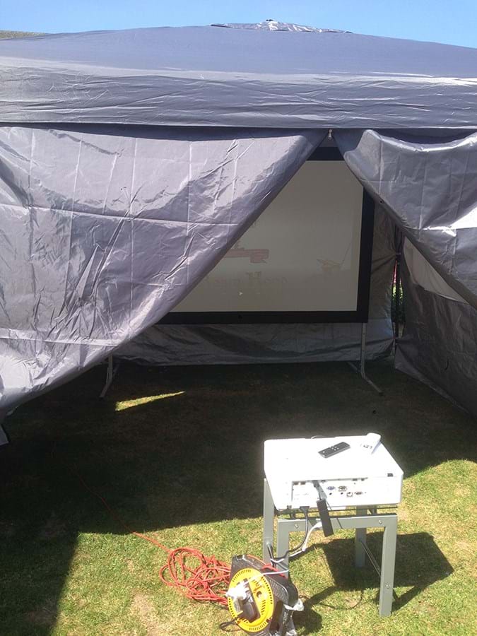 A film projector on a low table, pointed at a screen in a tent 
