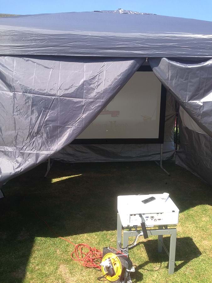 A film projector on a low table, pointed at a screen in a tent 