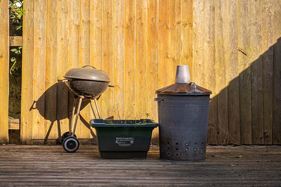 A barbecue, green plastic box and a bin against a wooden fence 