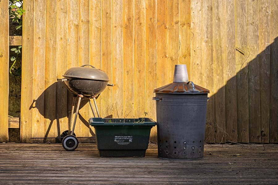 A barbecue, green plastic box and a bin against a wooden fence