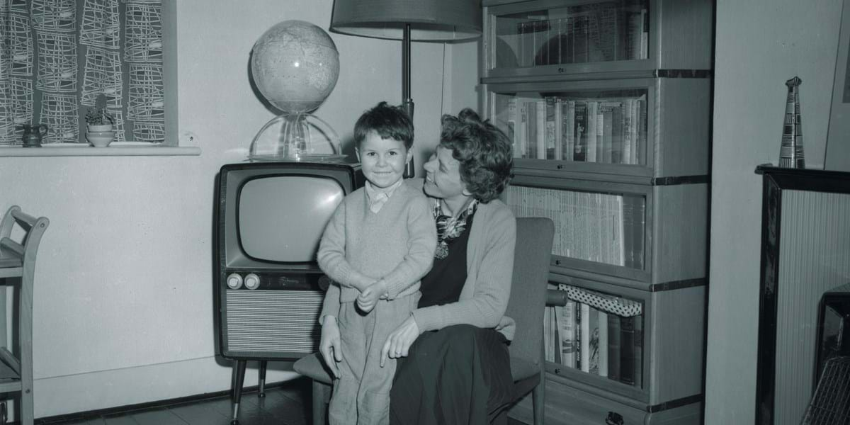 Black and white image of a child standing with an adult crouching next to them, in front of a television set and a bookshelf.