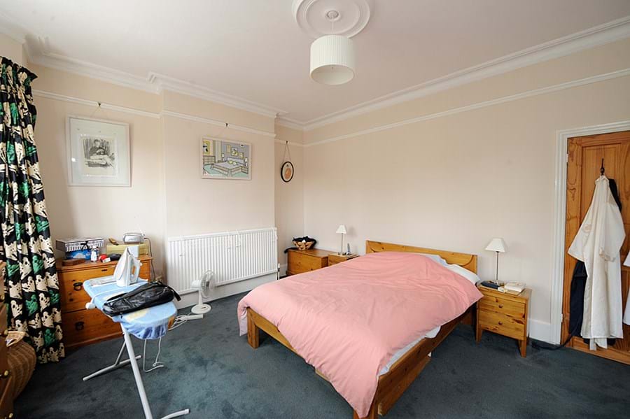 Photo of a bedroom with a bed with pink duvet, radiator on the wall and ironing board set up