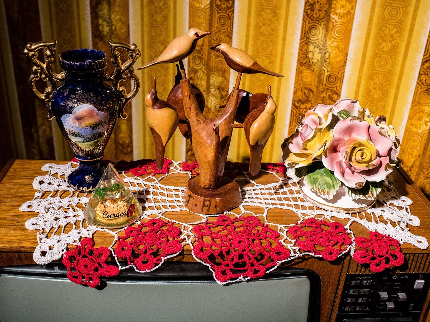 Old television set topped with various ornaments including red and white crochet and some wooden birds