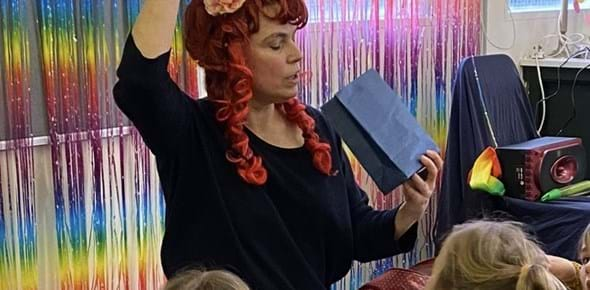 A person with a red wig on performing in front of a colourful backdrop to a group of children looking at them 