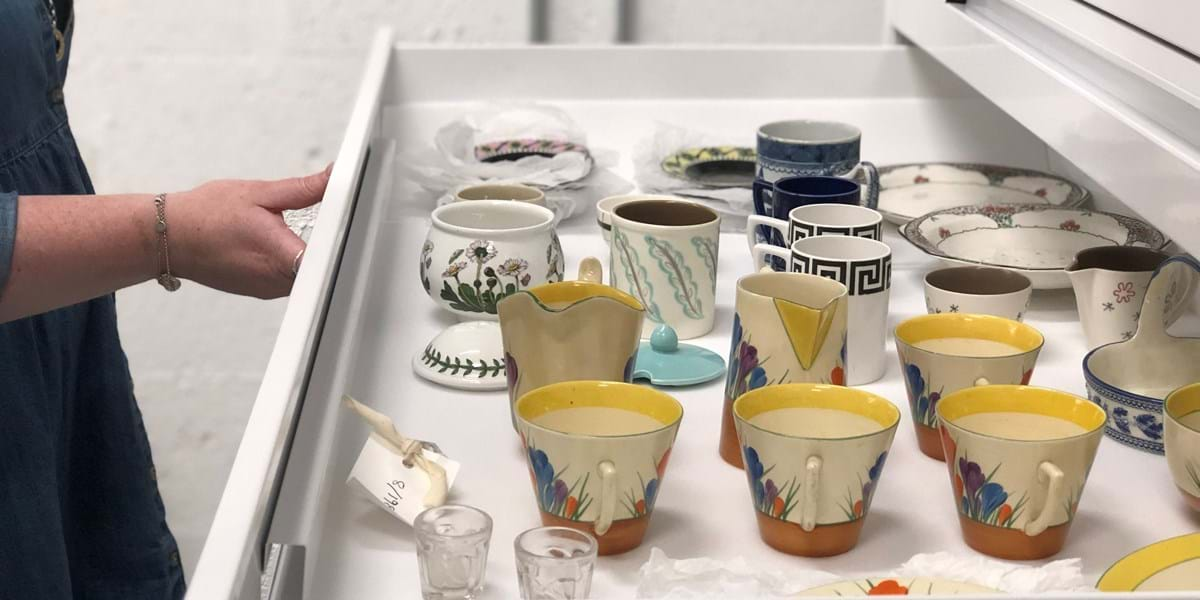 A person holding open a drawer in the Museum stores to show a range of colourful cups and glasses 