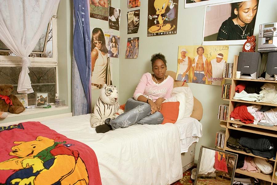 A young person leaning on pillows on a bed, with CDs and clothes on nearby shelves and posters on the walls