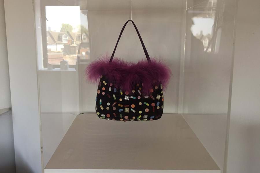 Black handbag with pills stitched in and purple feathers, displayed in a glass cabinet