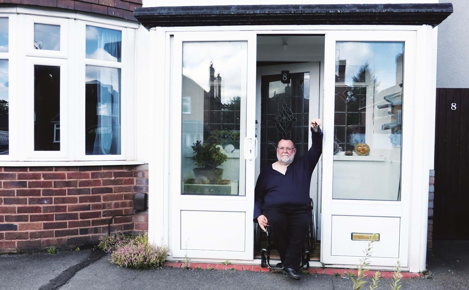Photo of a person sitting in a wheelchair in between their porch door, with left hand leant against the door frame