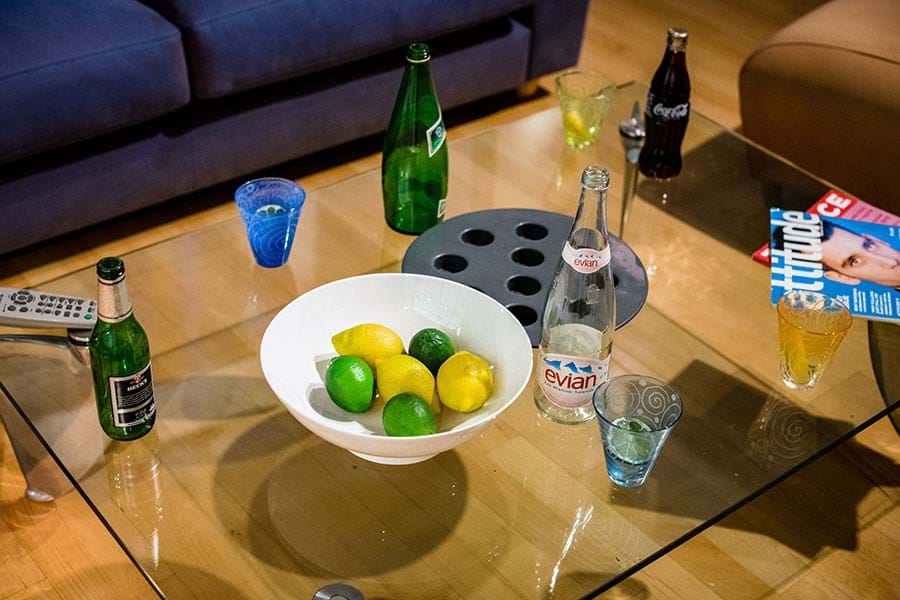 A glass table set with a white bowl of lemons and limes, various glass bottles and magazine