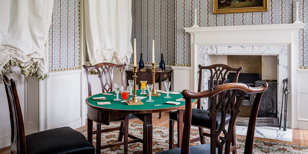 Wallpapered room with green card table and chairs set up for a game of cards, fireplace in background