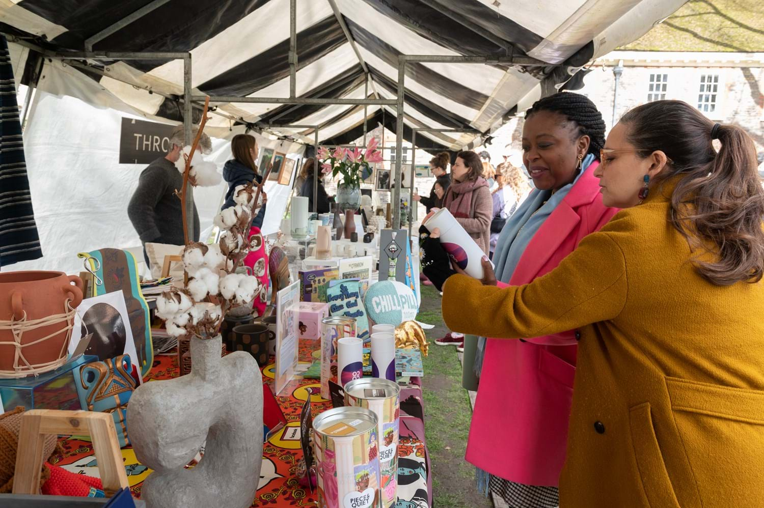 Two people curiously looking at an item at a yard sale, in front to a table full of colourful interior decoration items under a gazebo