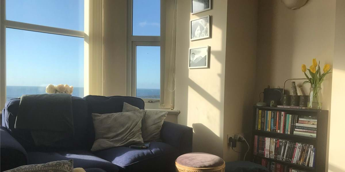 A view of the sea out of the window with a sofa and shelving unit in the foreground