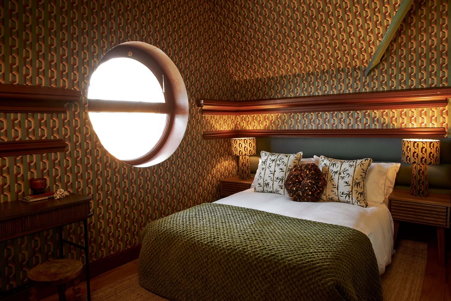 A bedroom decorated with geometric wallpaper in natural green tones with dark red accents, with a bed with matching lamps and pillows.
