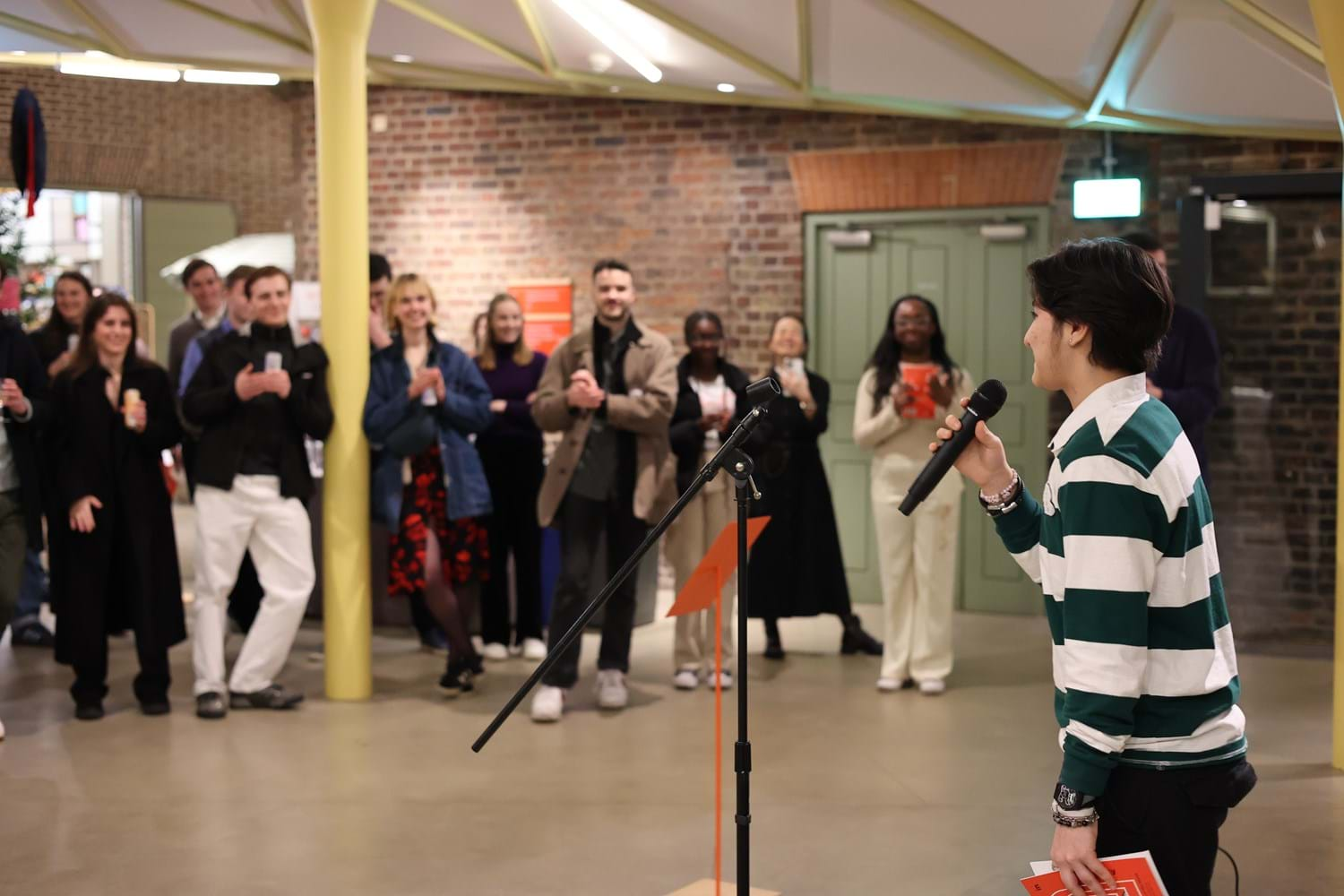 A person speaking into a microphone with an audience smiling at them while listening in the background