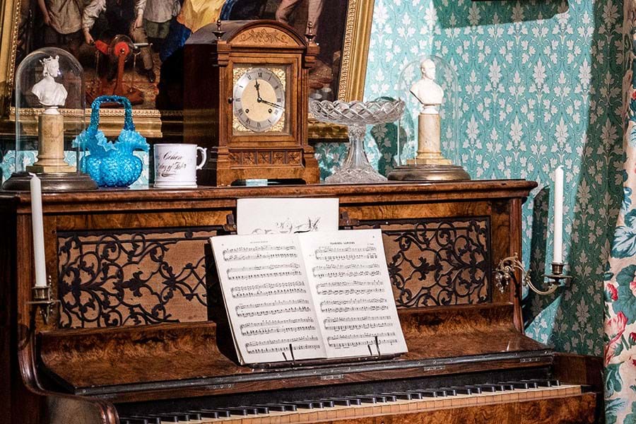 An open piano with a book of music set up. On top of the piano are various ornament including small busts under glass domes and a clock