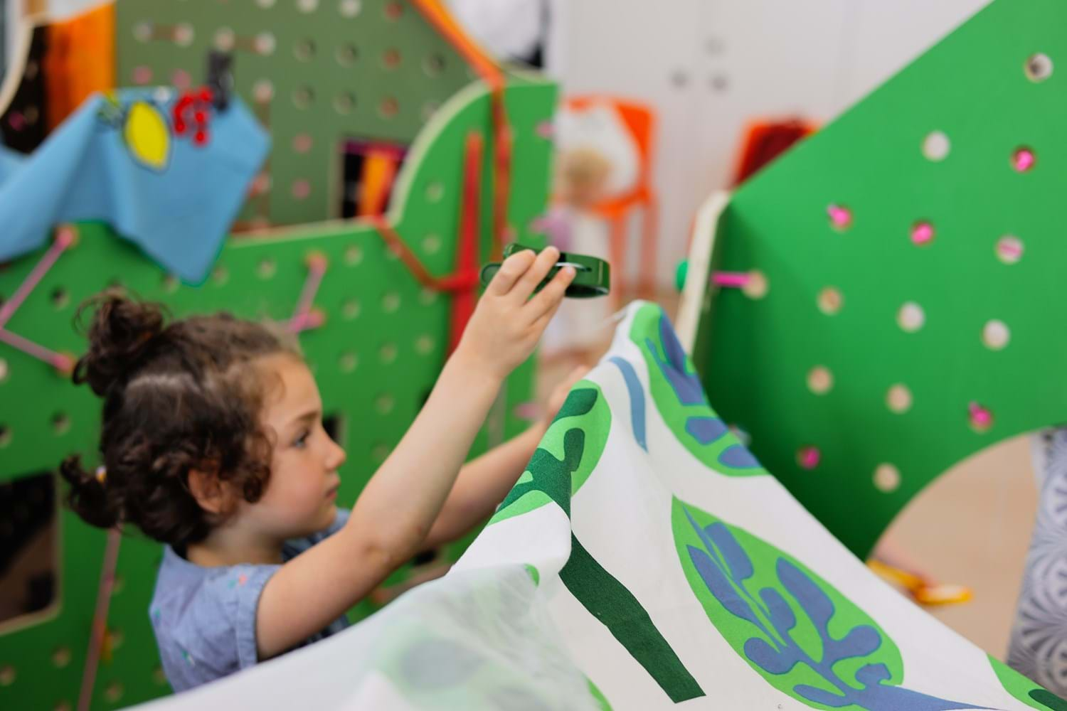 A child reaches up to clip a printed sheet of material over a wooden green structure.