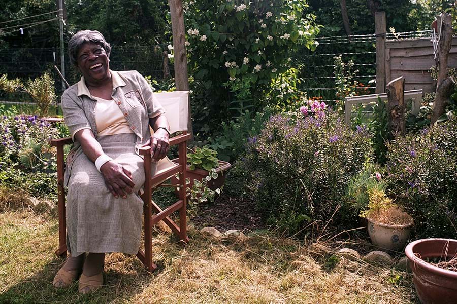A person laughing, they are seated on a wooden chair in a garden