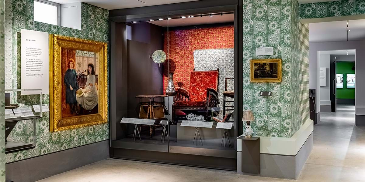 A museum gallery display of furniture and textiles in a glass cabinet, next to a painting hung on the wall 