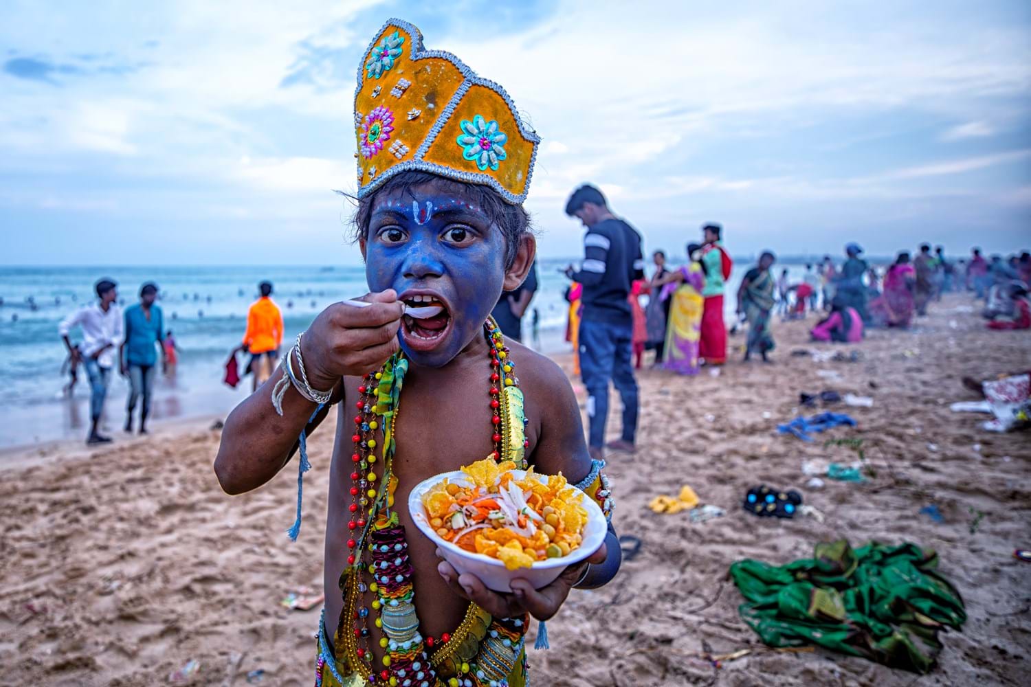 A child in India with a painted blue face, eating a bowl of food on a beach 