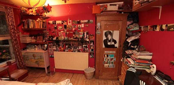 Colour photograph of a bedroom with red walls and posters and photos on the wall