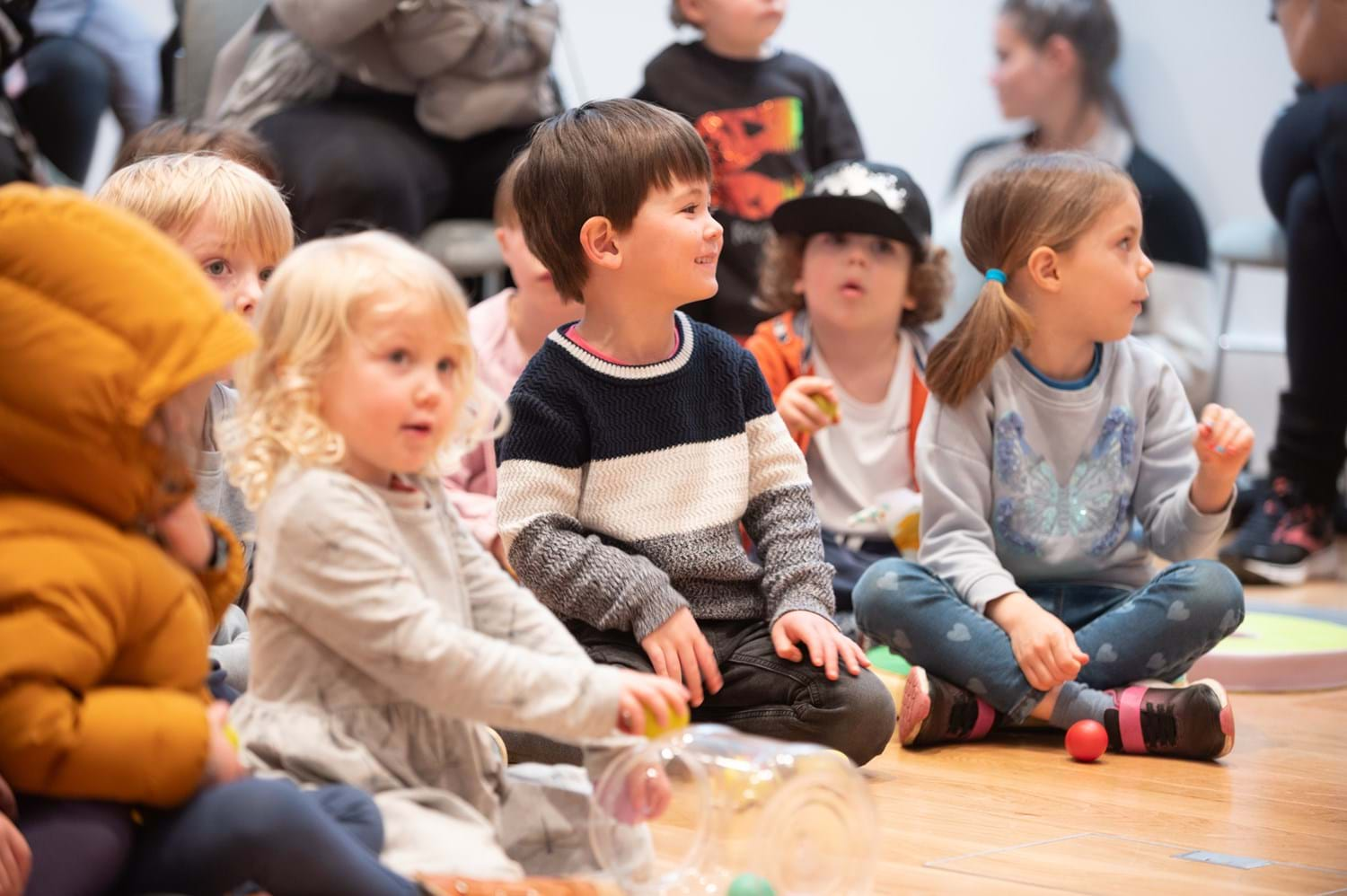 A group of children sitting cross legged on the floor smiling and listening to someone speak