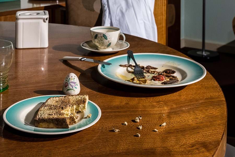 A recently abandoned breakfast table. Two blue plates, one has half eaten toast on it and the other has the remains of a fry up. There are crumbs on the table as well as a pepper shaker