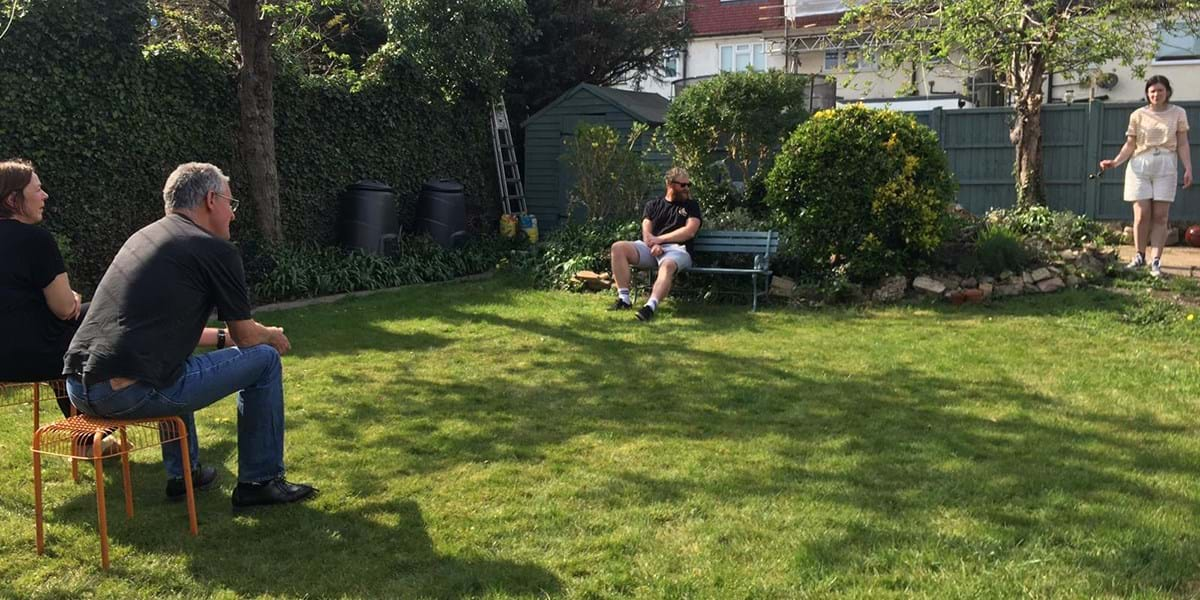 Four people socially distancing in a garden, two on stools, one on a bench and one standing