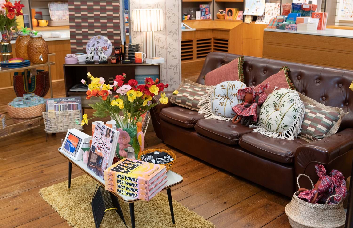 A brown leather sofa with floral throw pillows in natural tones surrounded by items from a museum shop on display 