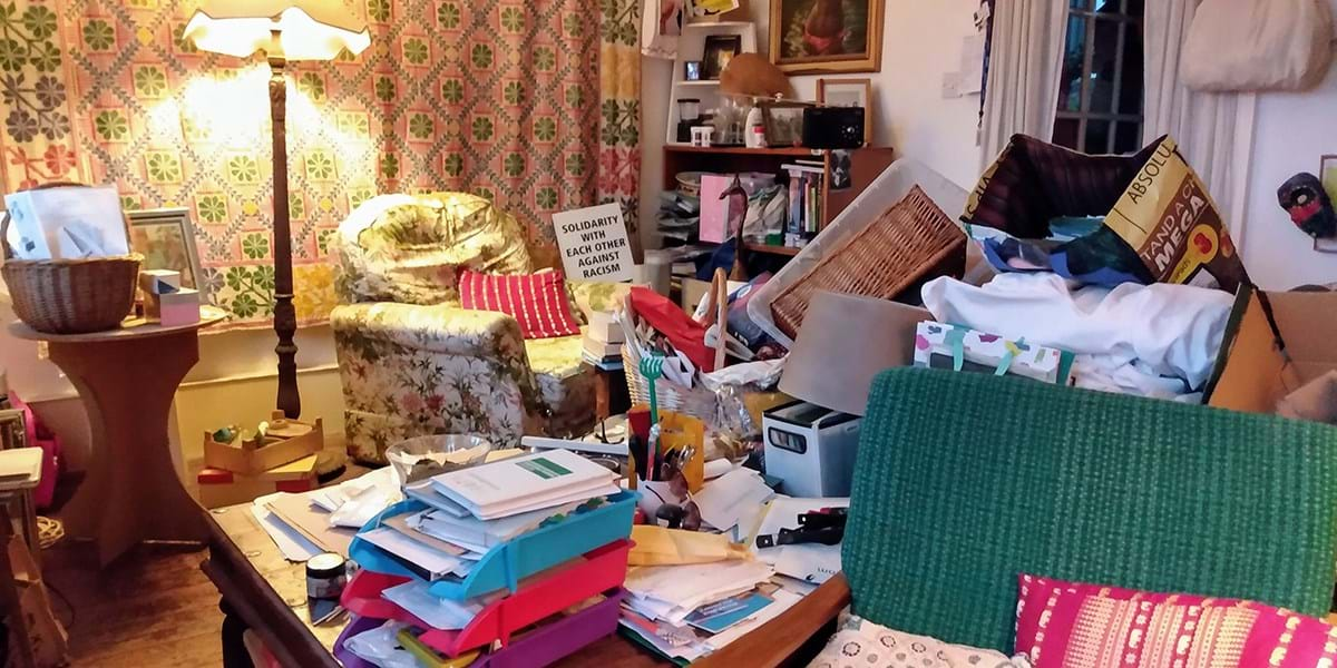 A living room with piles of paper, items of furniture and pictures on the walls 