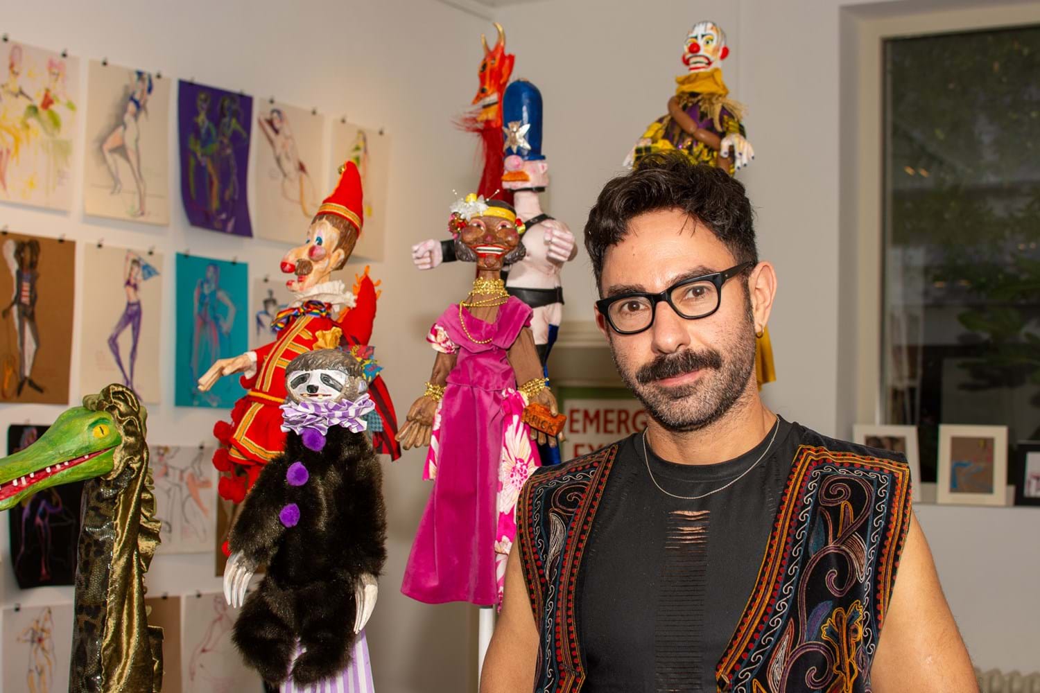 Oliver Hymans wearing black thick rimmed glasses standing in front of several colourful handmade puppets