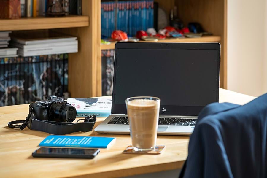 A camera, cup of coffee and a laptop on a desk in front of shelves of books