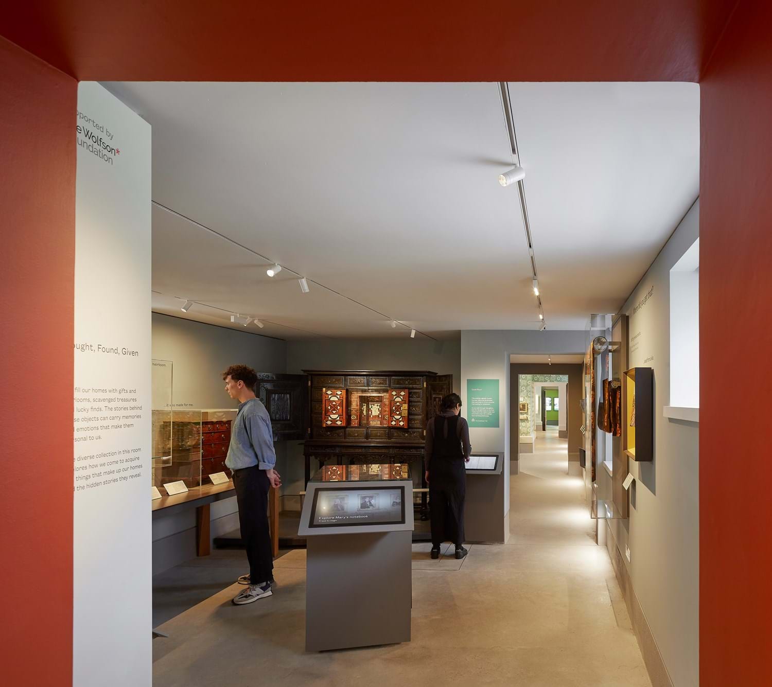 A long museum gallery with spaces to the left displaying furniture, interactive screens and glass cases