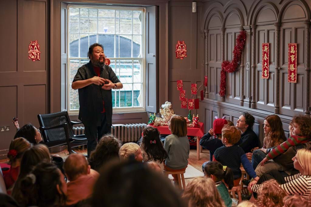 A person storytelling to a group of young children and some adults sitting on the floor, red lunar new year decorations are in the background