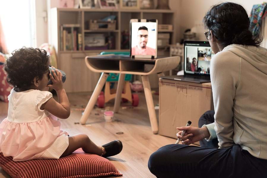 A small child sat on a cushion next to a person facing a television screen doing a video call
