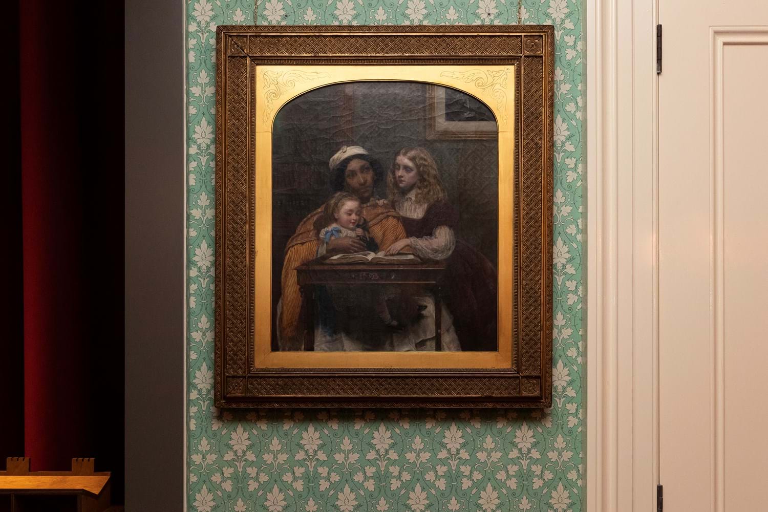 A painting by Rebecca Solomon, A Young Teacher, hanging on a wall depicting a woman tutoring two young children