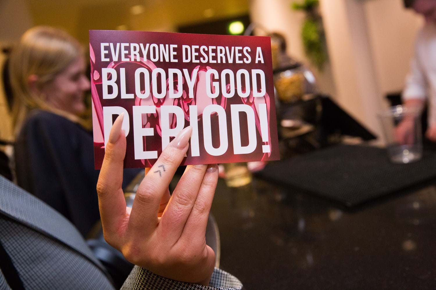 A hand holds a card to the camera reading Everyone Deserves a Bloody Good Period