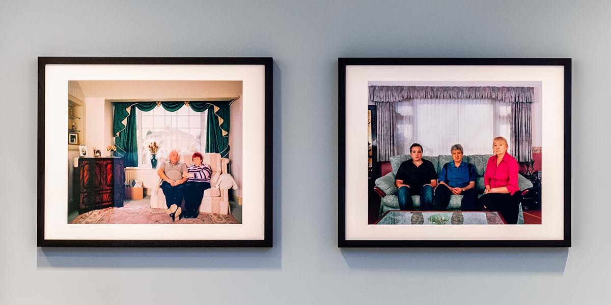 Two photos hung on a grey wall. Each photo depicts families sitting in their living rooms.