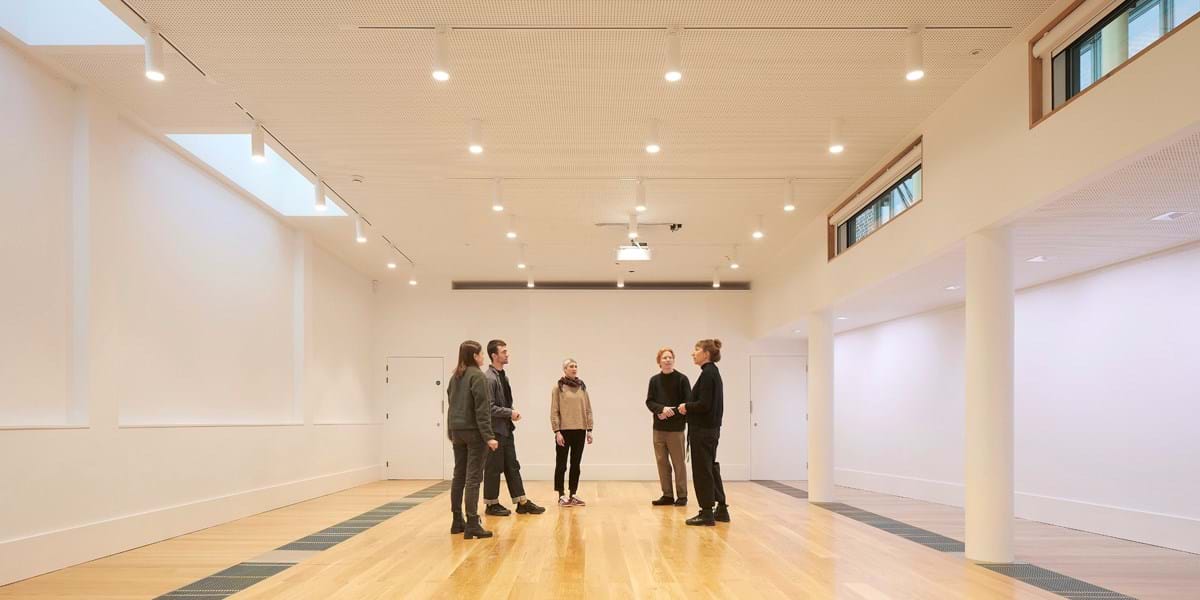 A group of people standing in a room with wooden floors, white walls and large windows
